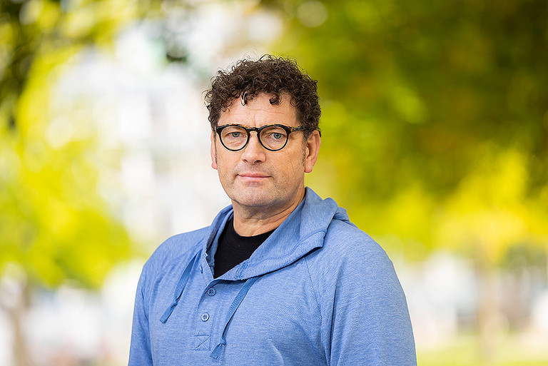 Das Foto zeigt das Portrait eines mittelalten Mann mit dunklen, kurzen lockigen haaren, einer dunklen Brille. Er trägt einen hellblauen Pullover und darunter ein schwarzes Tshirt.