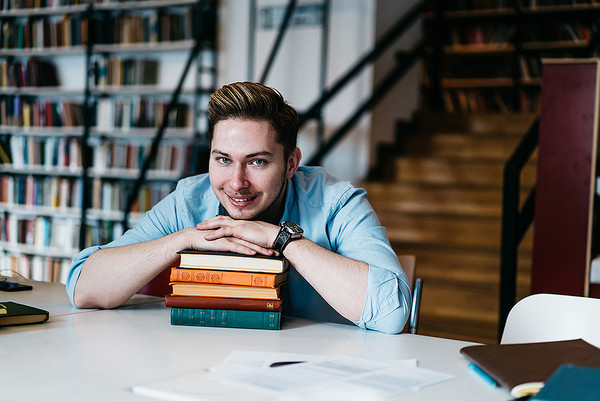 Ein junger Mann legt in einer Bibliothek sein Kinn auf einen Stapel Bücher