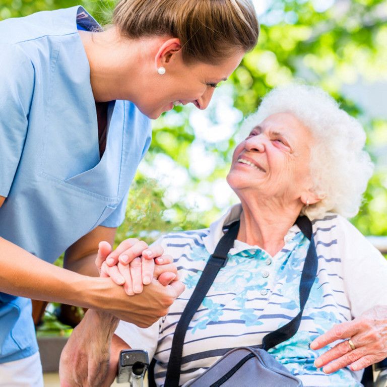 Pflegerin hält Seniorin im Rollstuhl aufmunternd die Hand