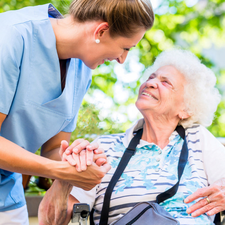 Pflegerin hält Seniorin im Rollstuhl aufmunternd die Hand