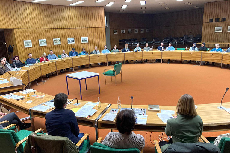 Viele Personen sitzen an Tischen in einem Kreis in einem Sitzungsraum.