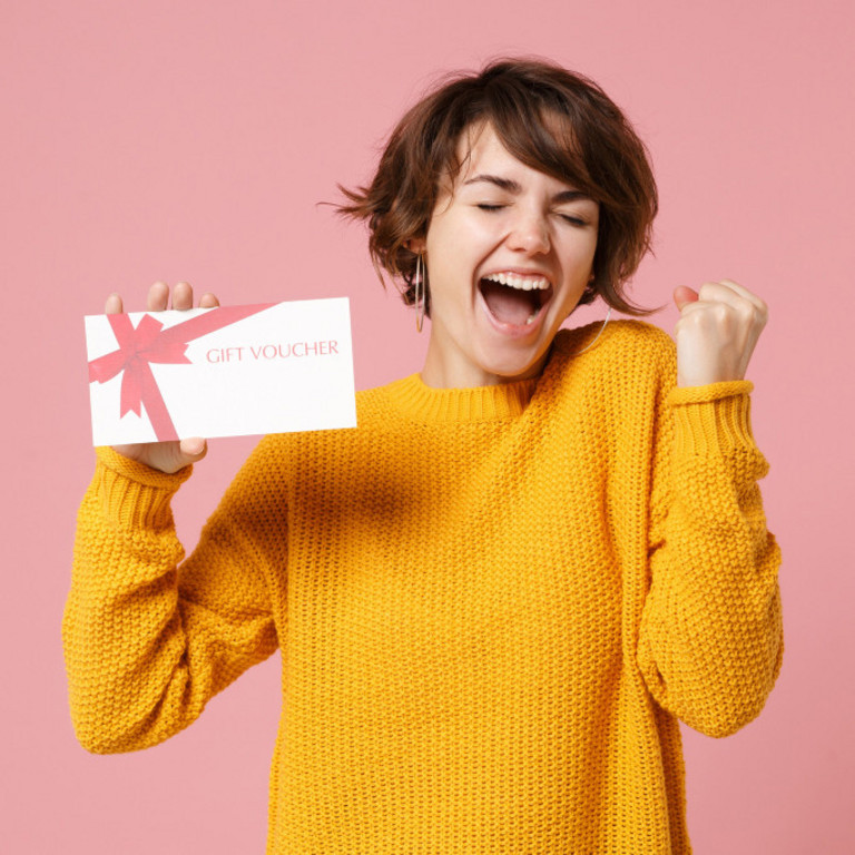 Fröhliche junge brünette Frau im gelben Pullover posiert isoliert auf pastellrosa Hintergrund im Studioporträt. Hält einen Geschenkgutschein in der Hand und macht eine Siegergeste. Menschen-Lifestyle-Konzept. 