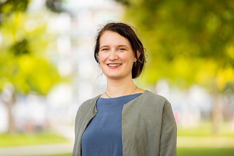 Das Foto ist ein Portrait einer jungen Frau mit dunklen, hochgesteckten Haaren, die in die Kamera lächelt. Sie trägt ein blaugraues Shirt und eine olivfarbene Jacke.