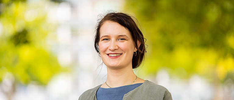 Das Foto ist ein Portrait einer jungen Frau mit dunklen, hochgesteckten Haaren, die in die Kamera lächelt. Sie trägt ein blaugraues Shirt und eine olivfarbene Jacke.