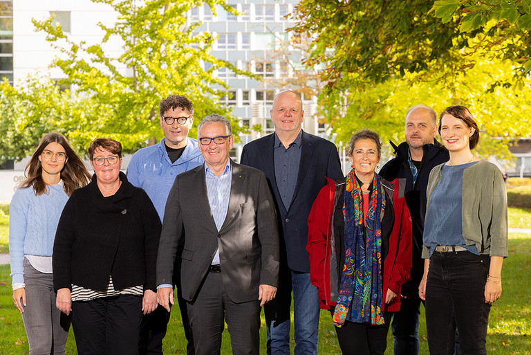 Das Foto zeigt eine Gruppe von lächelnden Menschen, die unter zwei Kastanienbäumen stehen. Ganz links steht eine junge Frau mit dunklen Haaren und blauem Pullover, daneben eine etwas ältere Frau im schwarzen Pullover, daneben ein gleichalter Mann in blauem Pullover, davor ein Mann mit grauen Harren und Sakko, dahinter ein ungefähr gleichalter Mann ebenfalls im Sakko, daneben eine Frau im gleichen Alter in roter Jacke, dahinter ein Mann in dunkler Jacke und daneben eine jüngere Frau mit grünem Blazer.