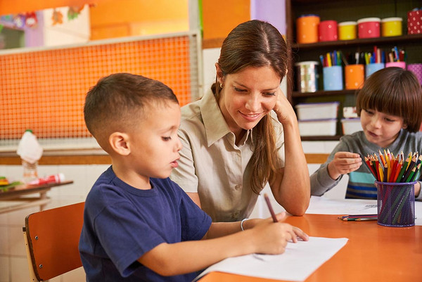 eine Kindergärtnerin sitzt mit einem Jungen der malt an einem Tisch