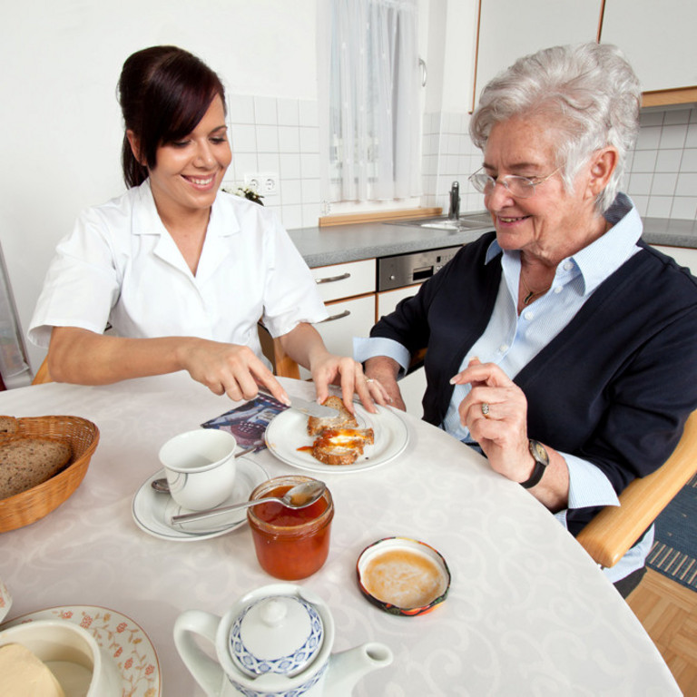 Mitarbeiterin eines Pflegedienstes hilft Dame beim Frühstück