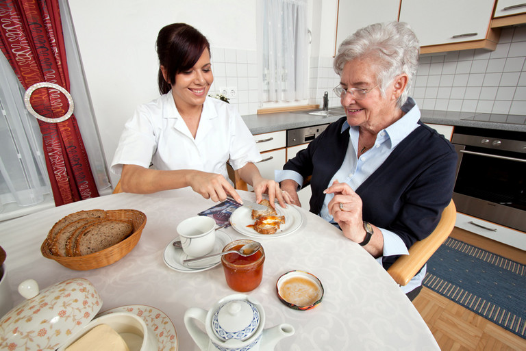 Mitarbeiterin eines Pflegedienstes hilft Dame beim Frühstück