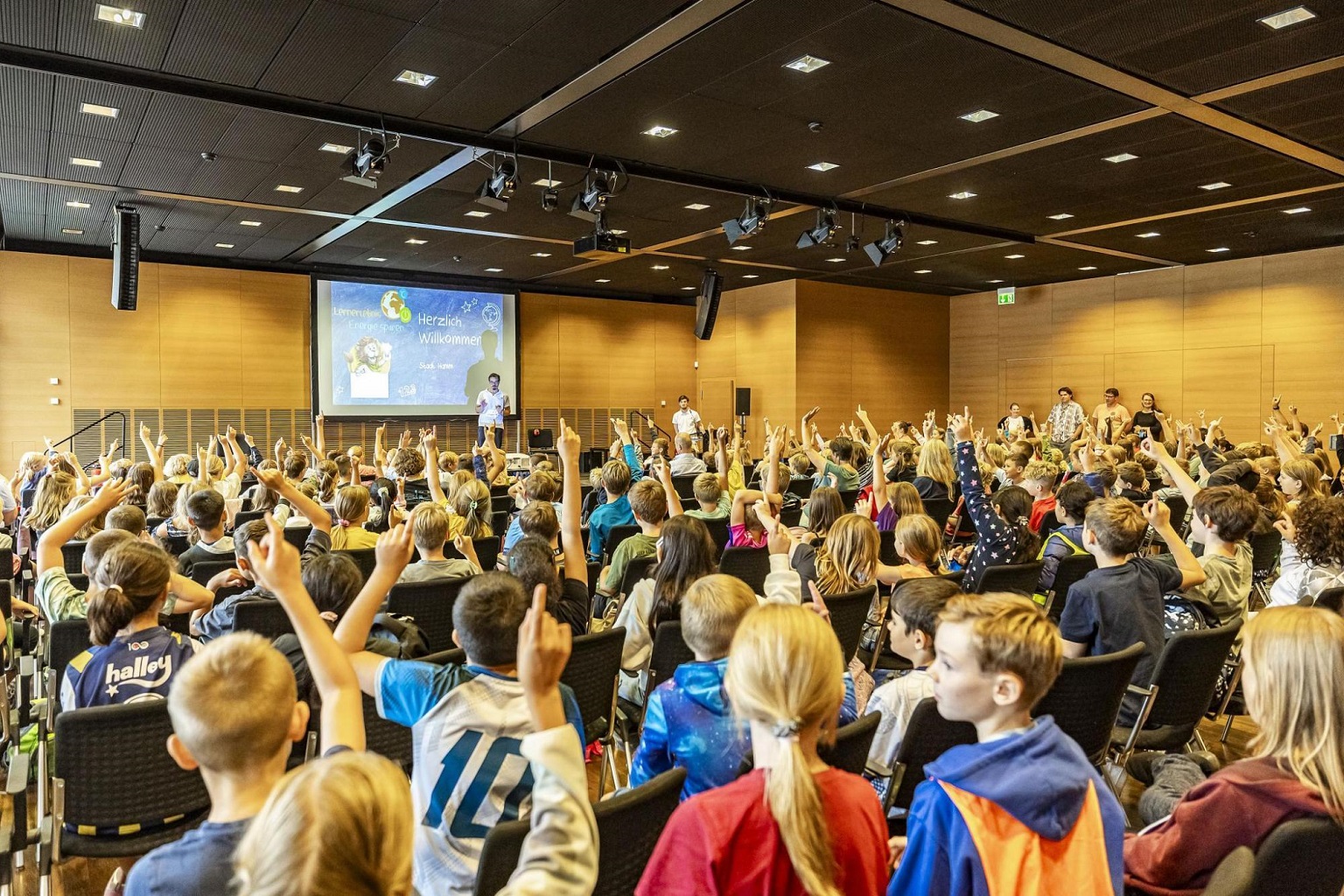 Das Foto zeigt einen voll besetzten Saal. Alle Kinder zeigen auf und nehmen am Quiz teil.