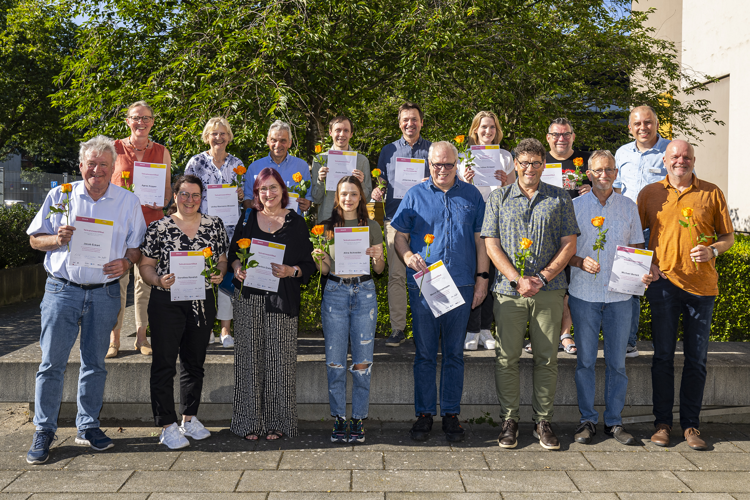 Das Foto zeigt die Menschen, die am Klima.fit Kurs 2024 teilgenommen haben. Sie stehen draußen, halten ihre Urkunden und eine gelbe Rose in der Hand und lächeln stolz in die Kamera.